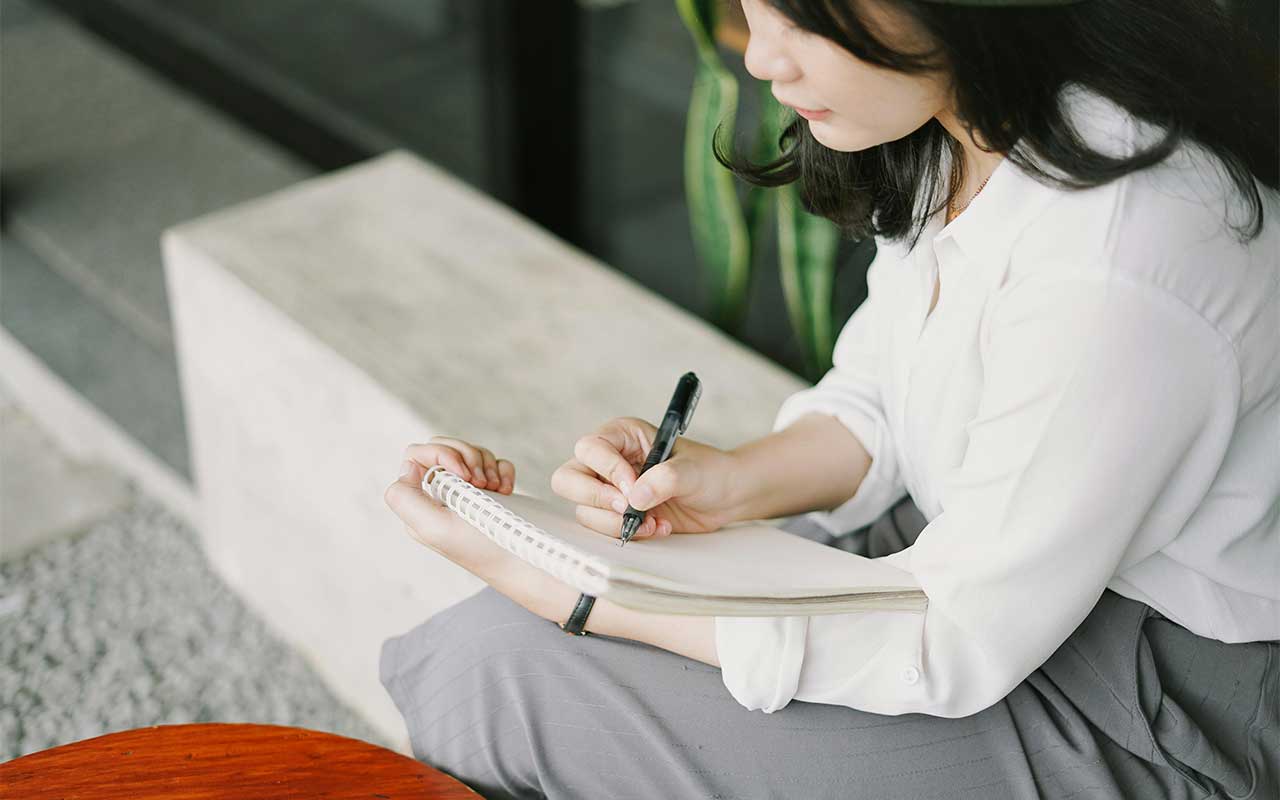 Woman writing a want list in a notebook during a no spend challenge to delay spending and stop impulse buys