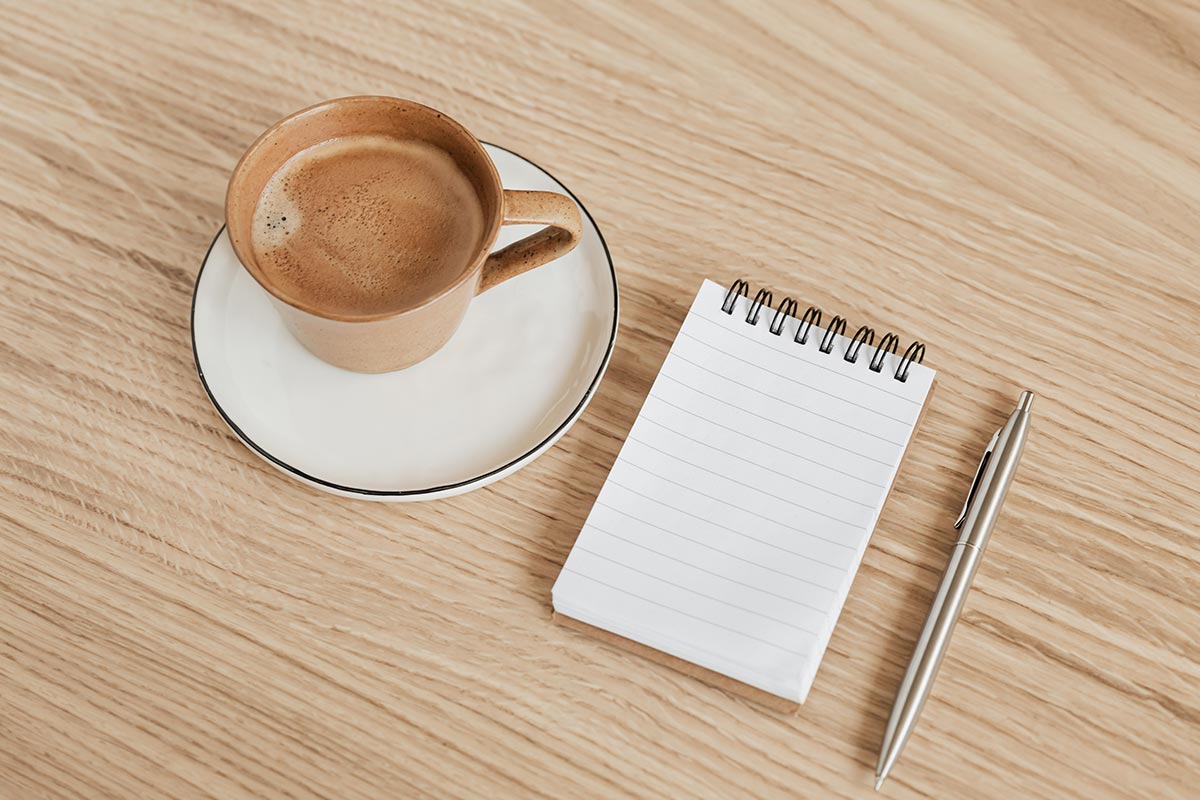 An open notebook, a pen, and coffee on a table — how to save money on groceries by planning before you shop
